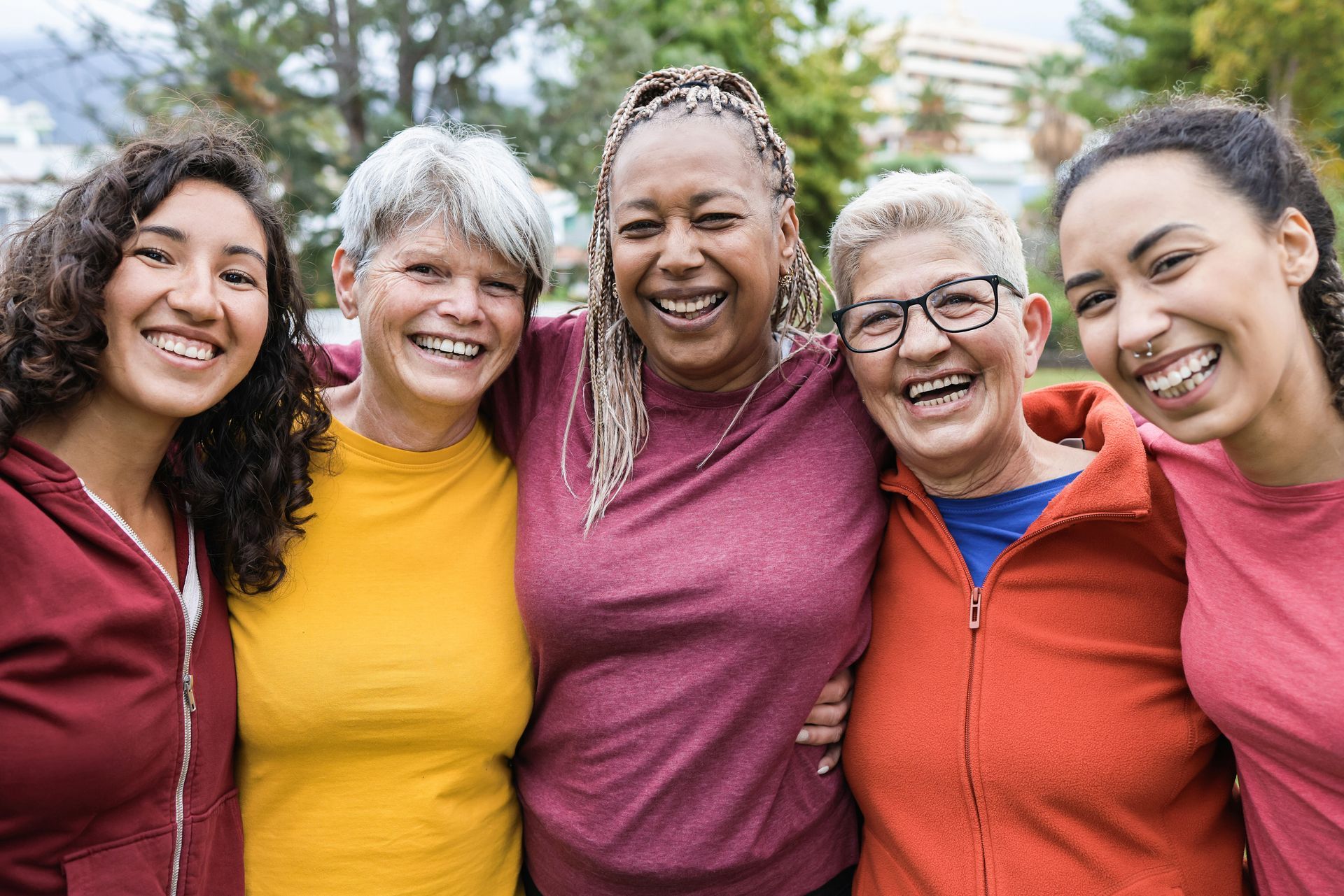 Five diverse women smiling with arms around each other outdoors. They are various ages with different hair and clothing colors.