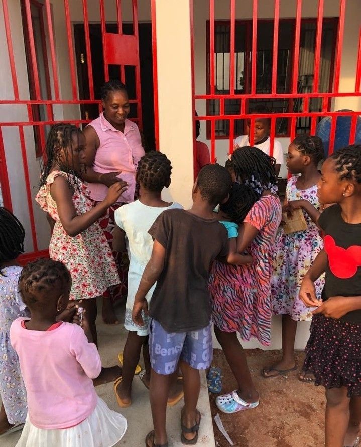A group of children gather around a woman in front of a building with red bars, laughing and playing.
