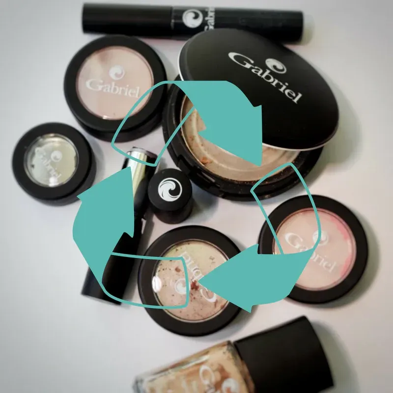 Makeup products arranged with a recycling symbol overlaid, suggesting recycling or eco-friendliness. Products are mostly black, with shades of pink and beige.