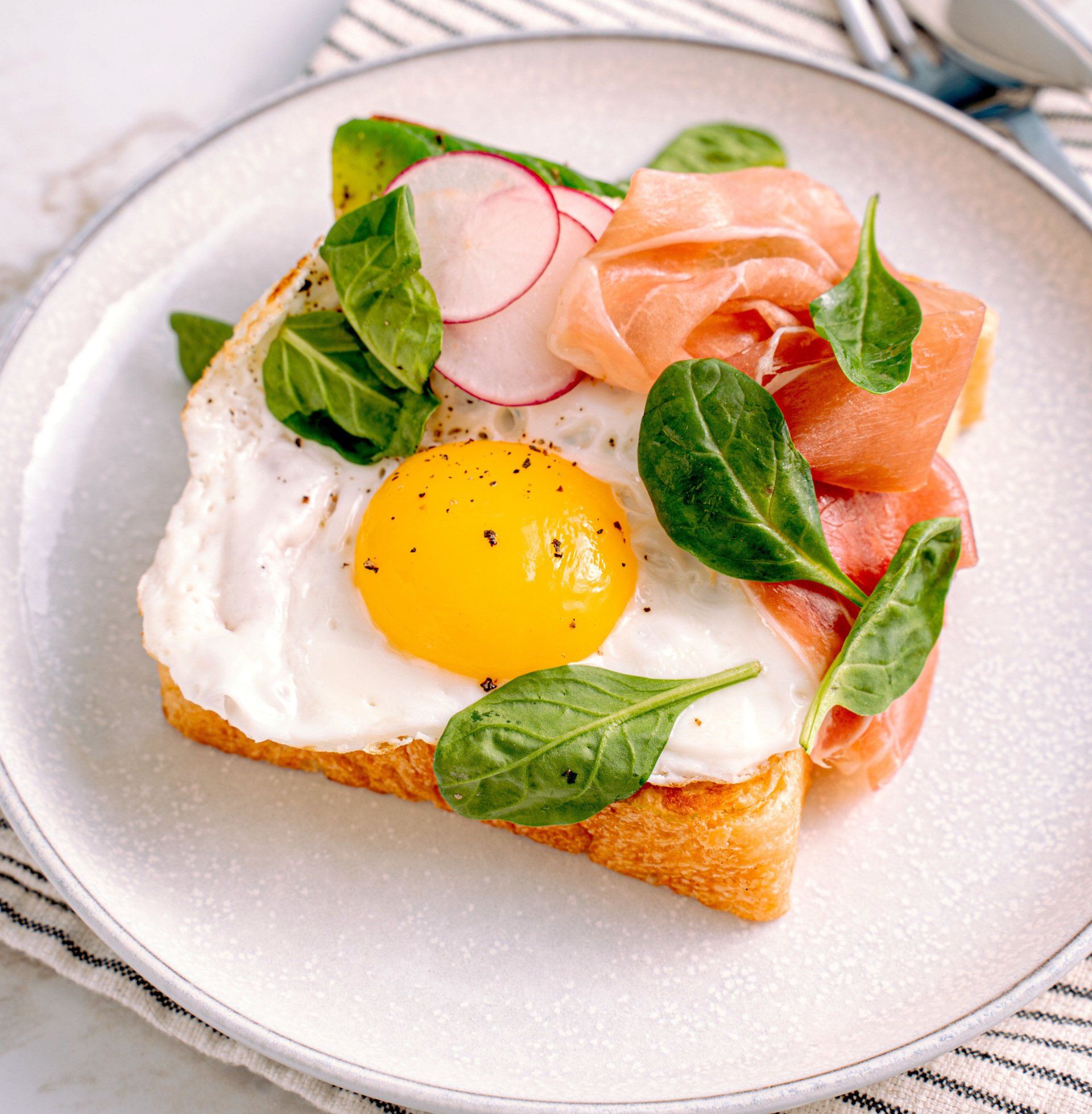 Fried egg on toast, topped with prosciutto, spinach, and radish slices on a white plate.