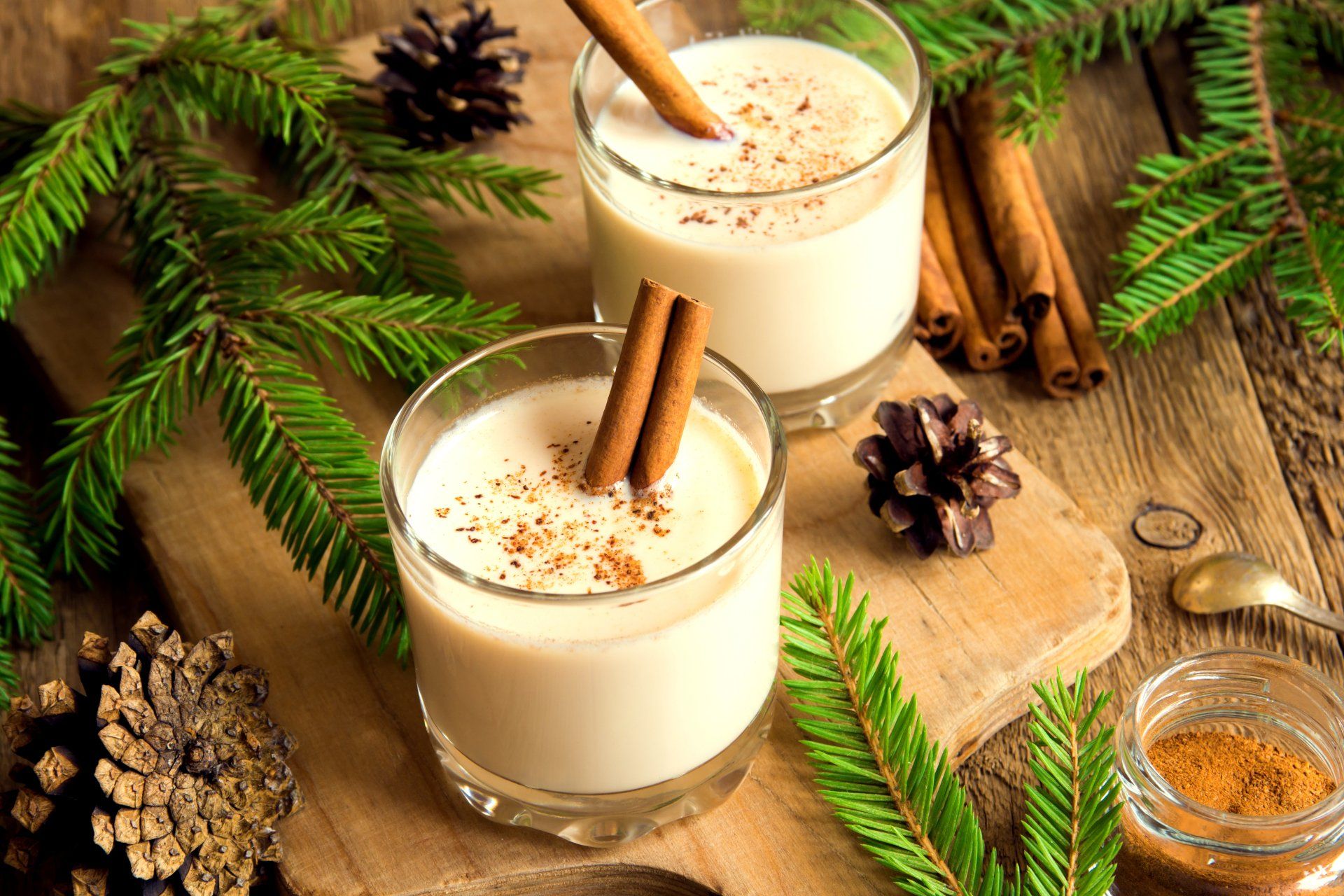 Two glasses of eggnog garnished with cinnamon sticks, set on a wooden board amidst pine branches and cinnamon sticks.
