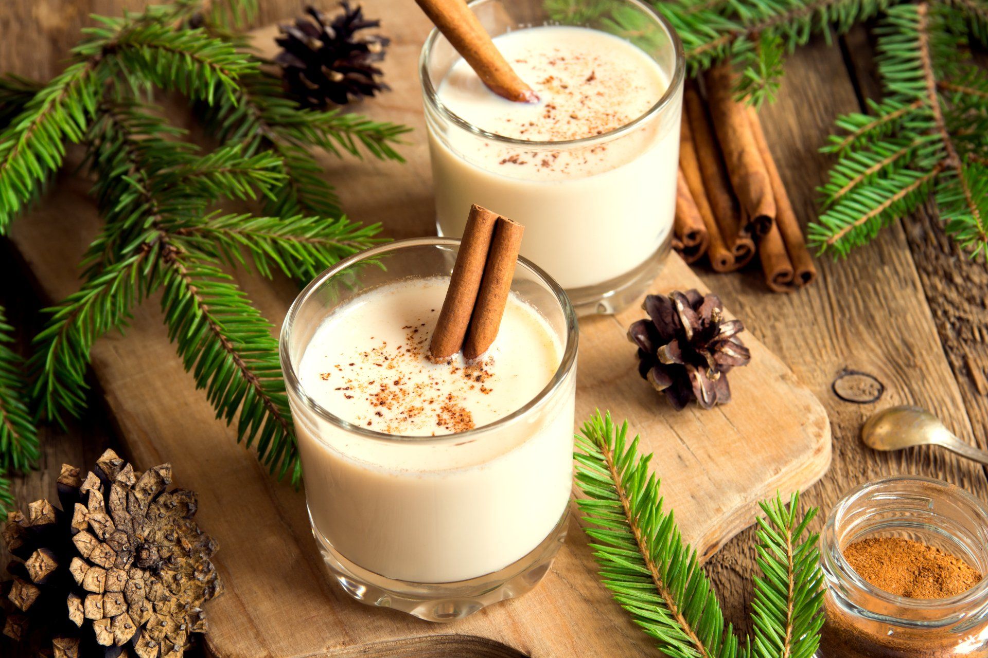 Two glasses of eggnog, garnished with cinnamon sticks and nutmeg, are arranged with pine boughs, pine cones, and cinnamon sticks on a wooden surface.