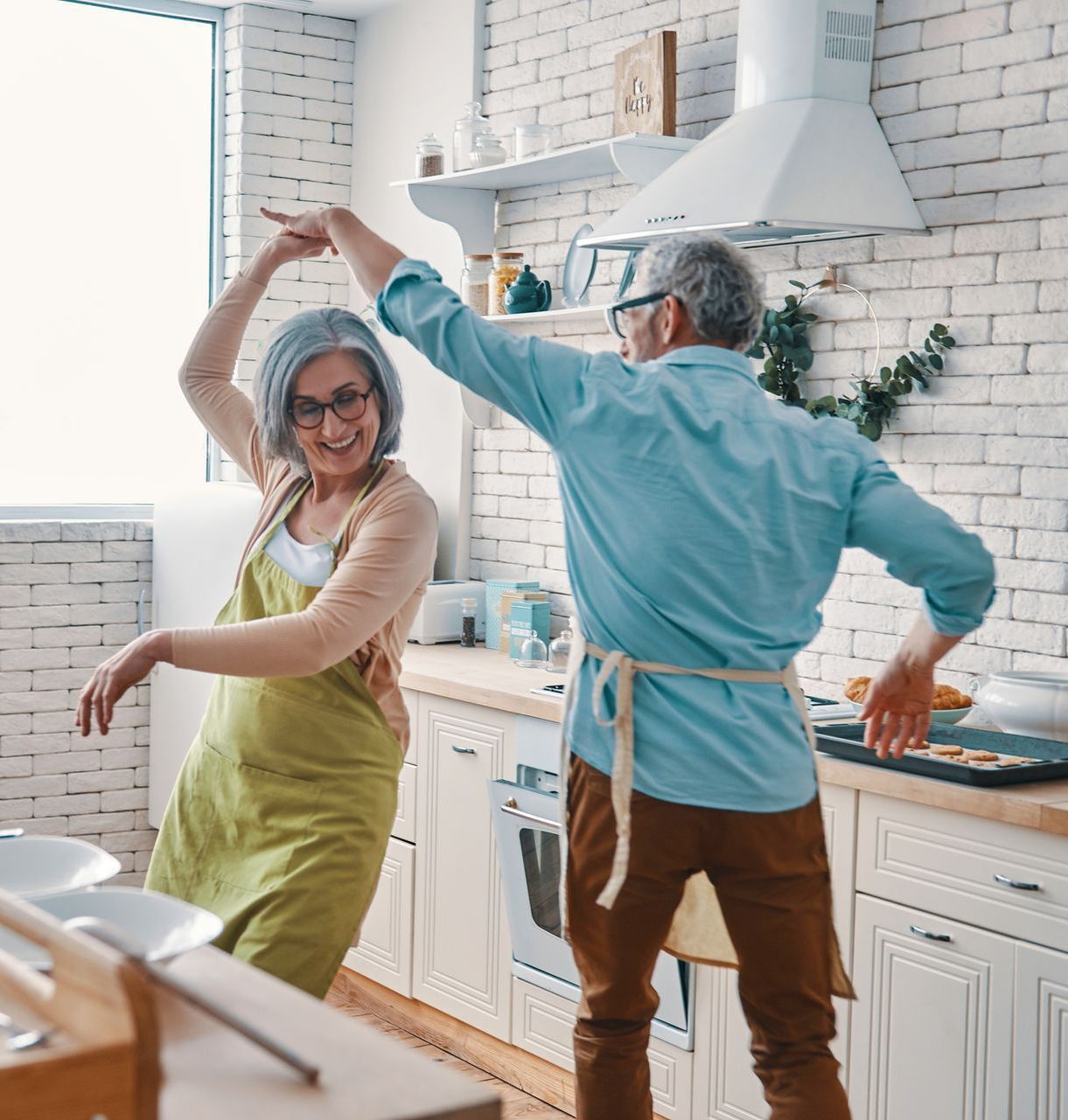 Elderly couple dancing joyfully in a bright kitchen; woman in apron, man in button-down.