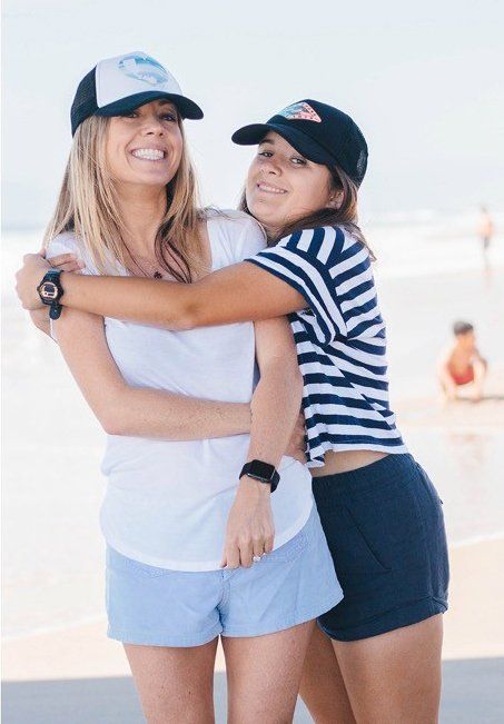 Two young women embrace on a beach, both wearing hats and watches. The one in front smiles, the other hugs her with a smile.