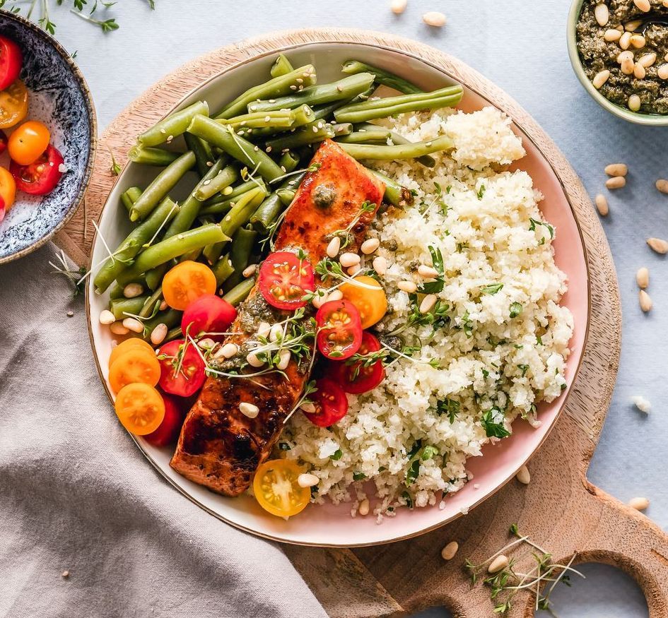 Salmon fillet, green beans, cauliflower rice, and tomatoes on a plate, set on a wooden board.
