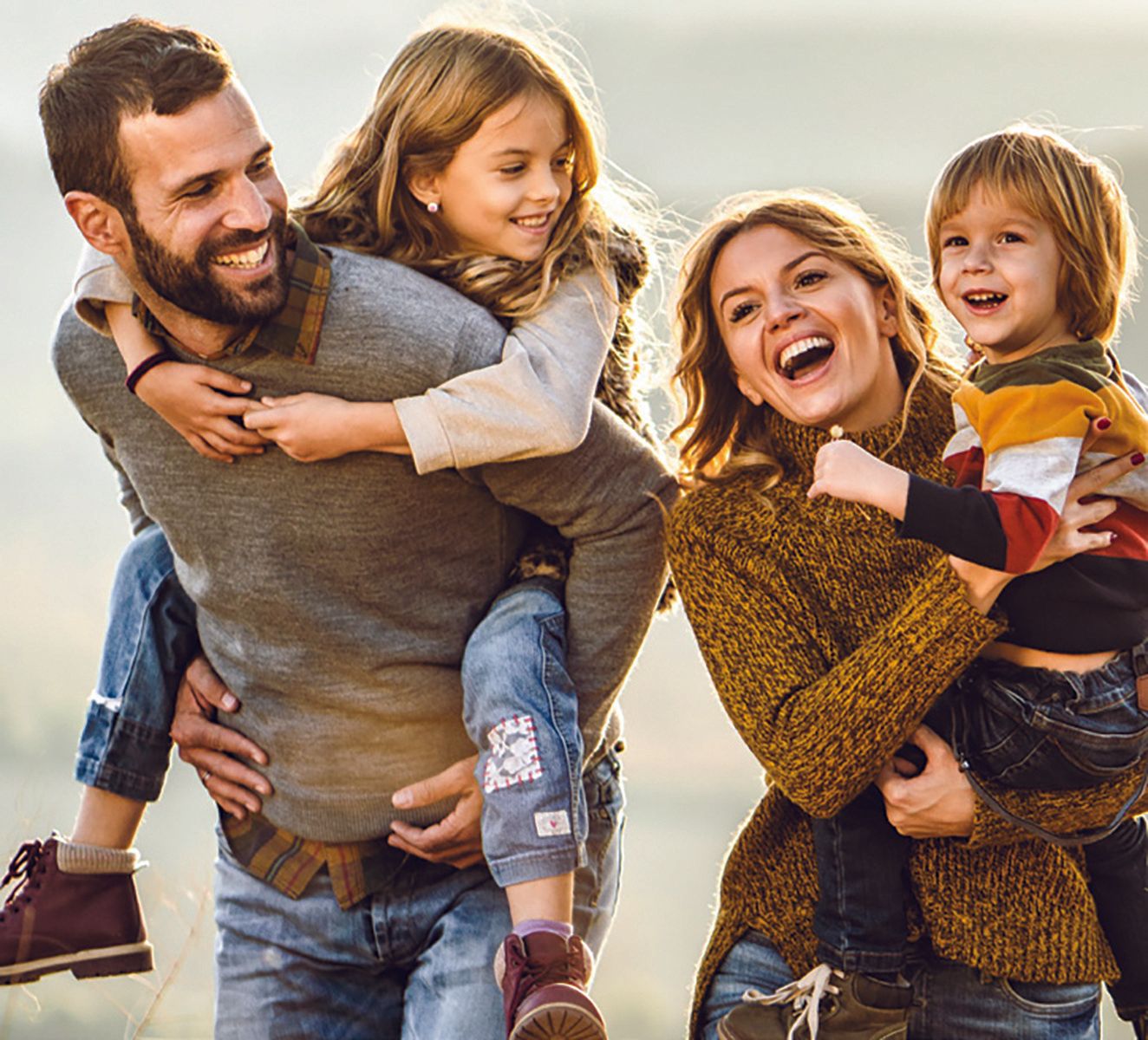 A happy family of four outdoors. A father gives a piggyback ride to a girl, and the mother holds a boy; all smiling.