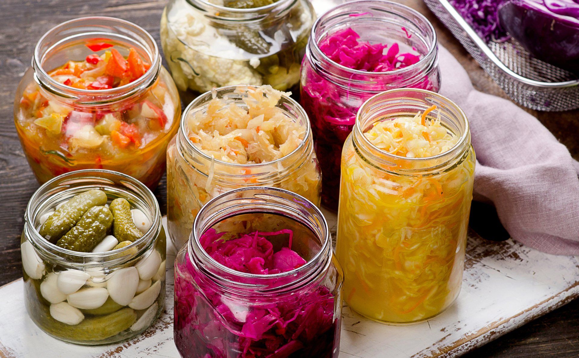 Jars of colorful fermented vegetables, including pickles, sauerkraut, and red cabbage, arranged on a wooden tray.