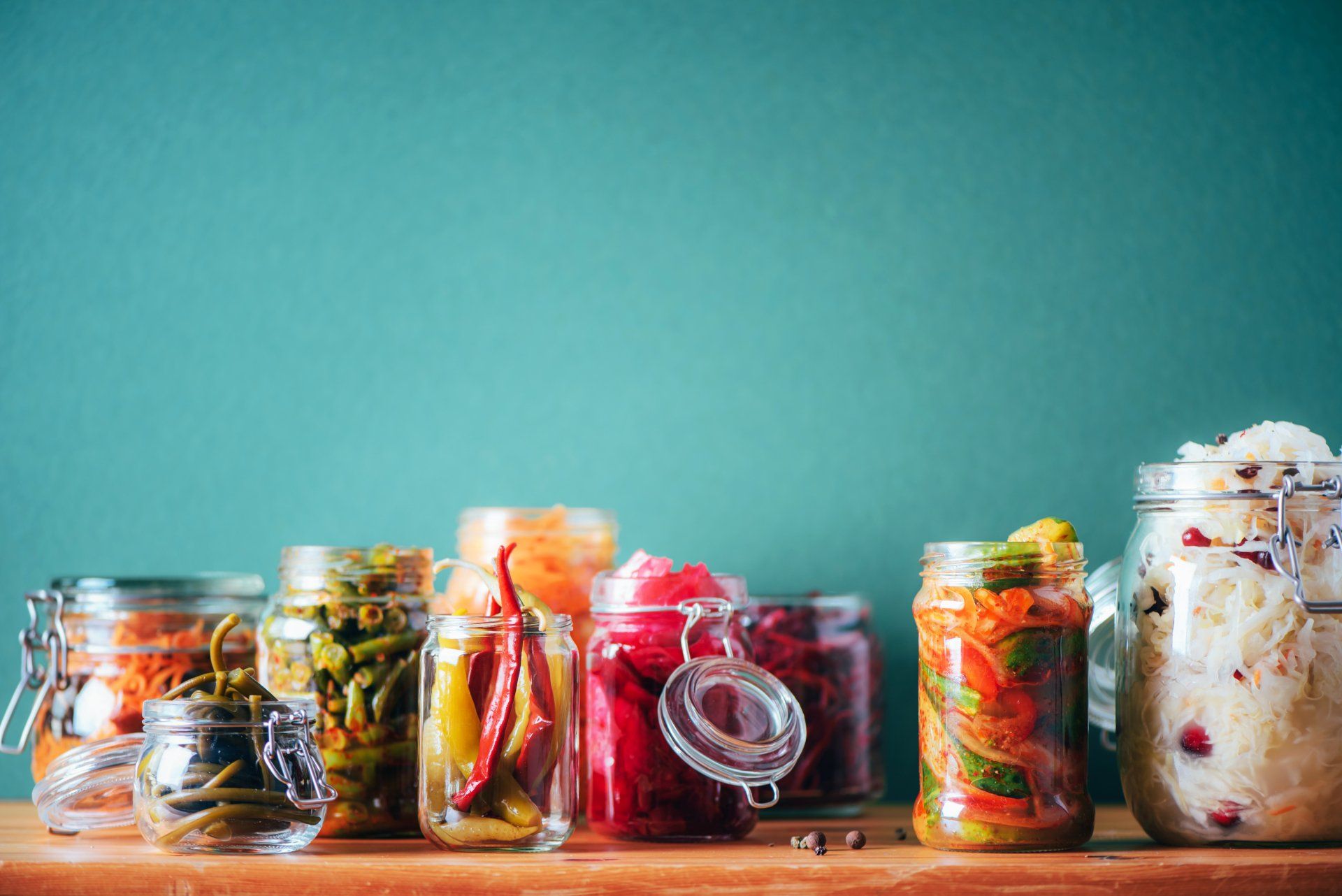 Jars of colorful fermented vegetables, including pickles and sauerkraut, on a wooden shelf against a teal background.
