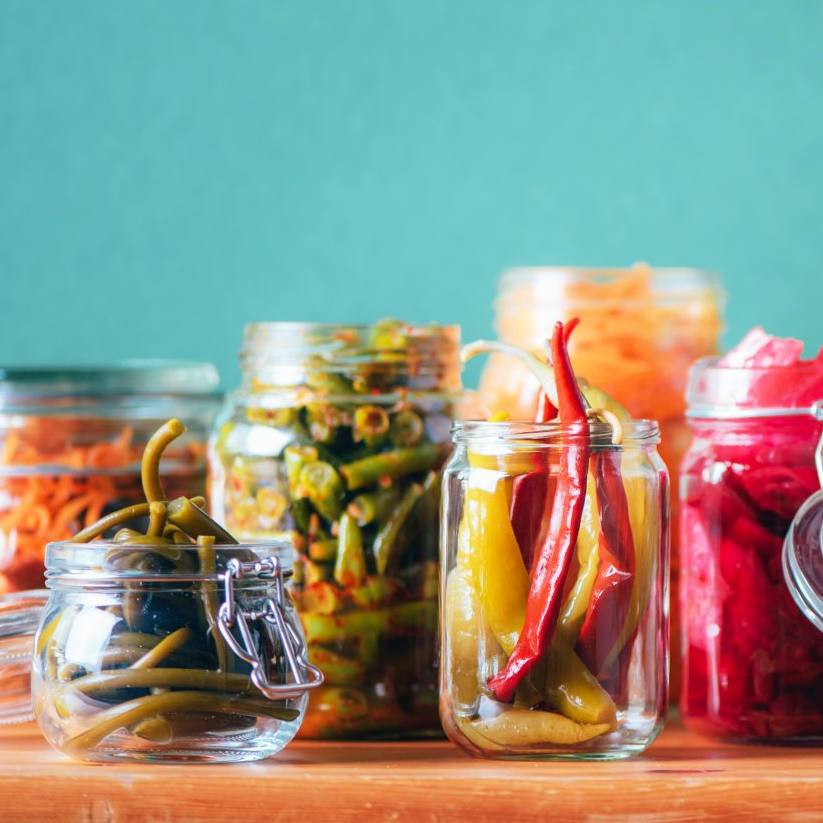 Assortment of colorful pickled vegetables in glass jars on a wooden surface against a teal background.