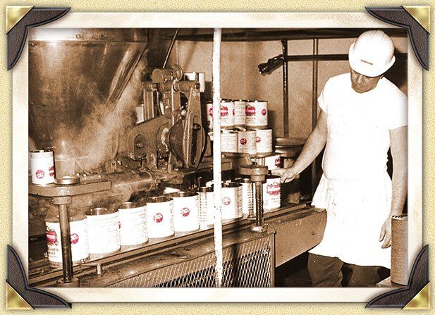 A vintage photo shows a man in a white uniform and hard hat on a factory line, overseeing the filling of cans.