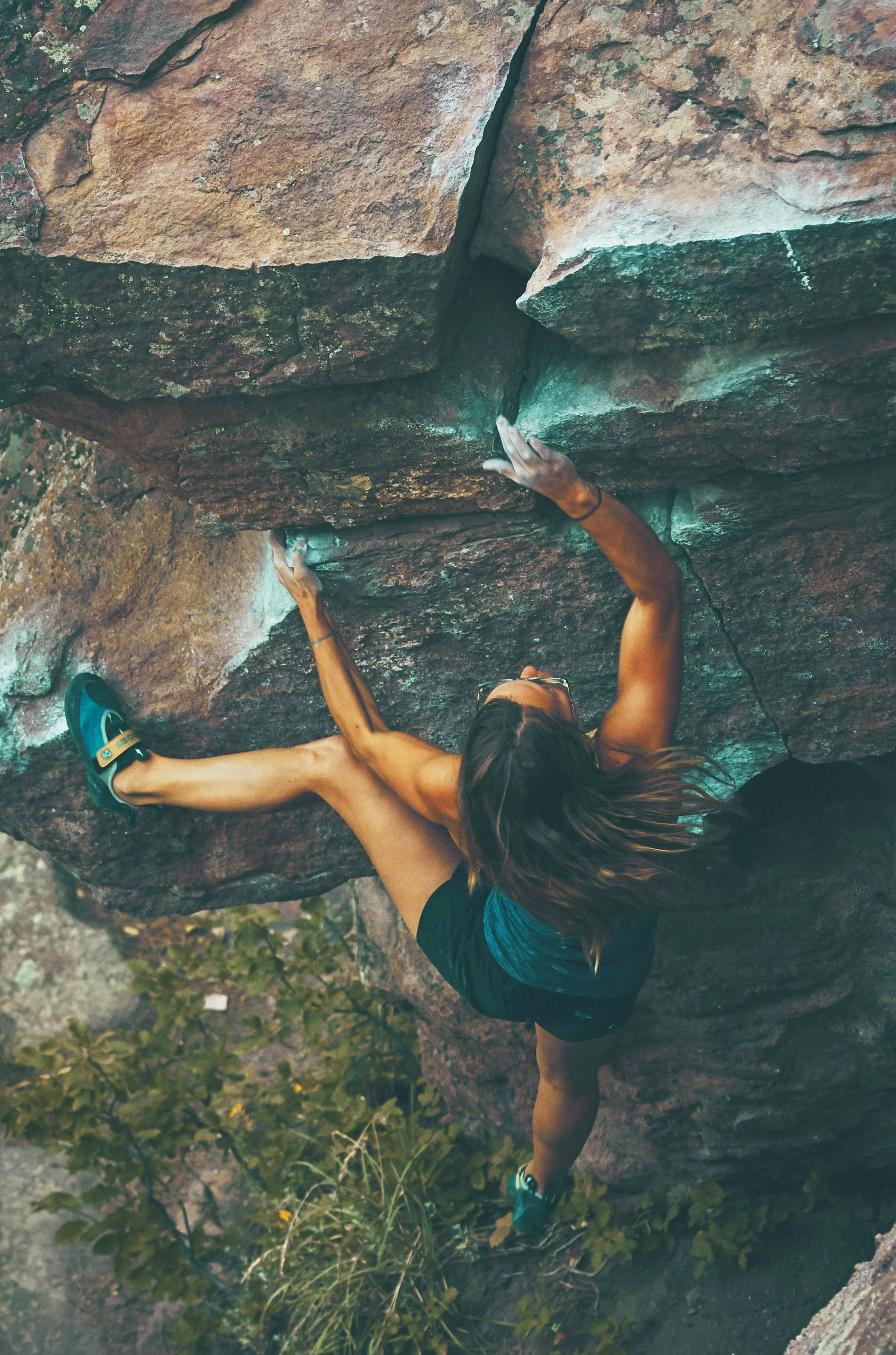 Woman rock climbing outdoors, reaching up for a hold on a textured rock face. She wears black shorts and is focused on the ascent.