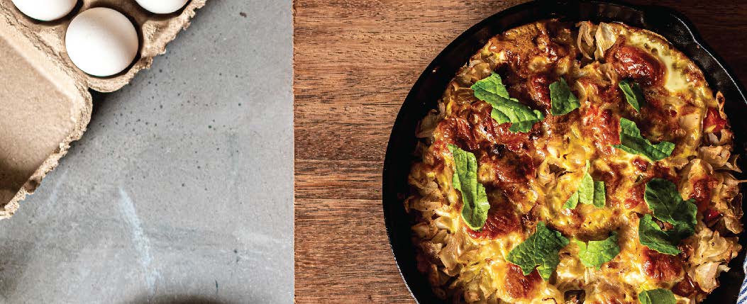 Eggs next to a skillet filled with frittata, garnished with green leaves, on a wood-grain surface.