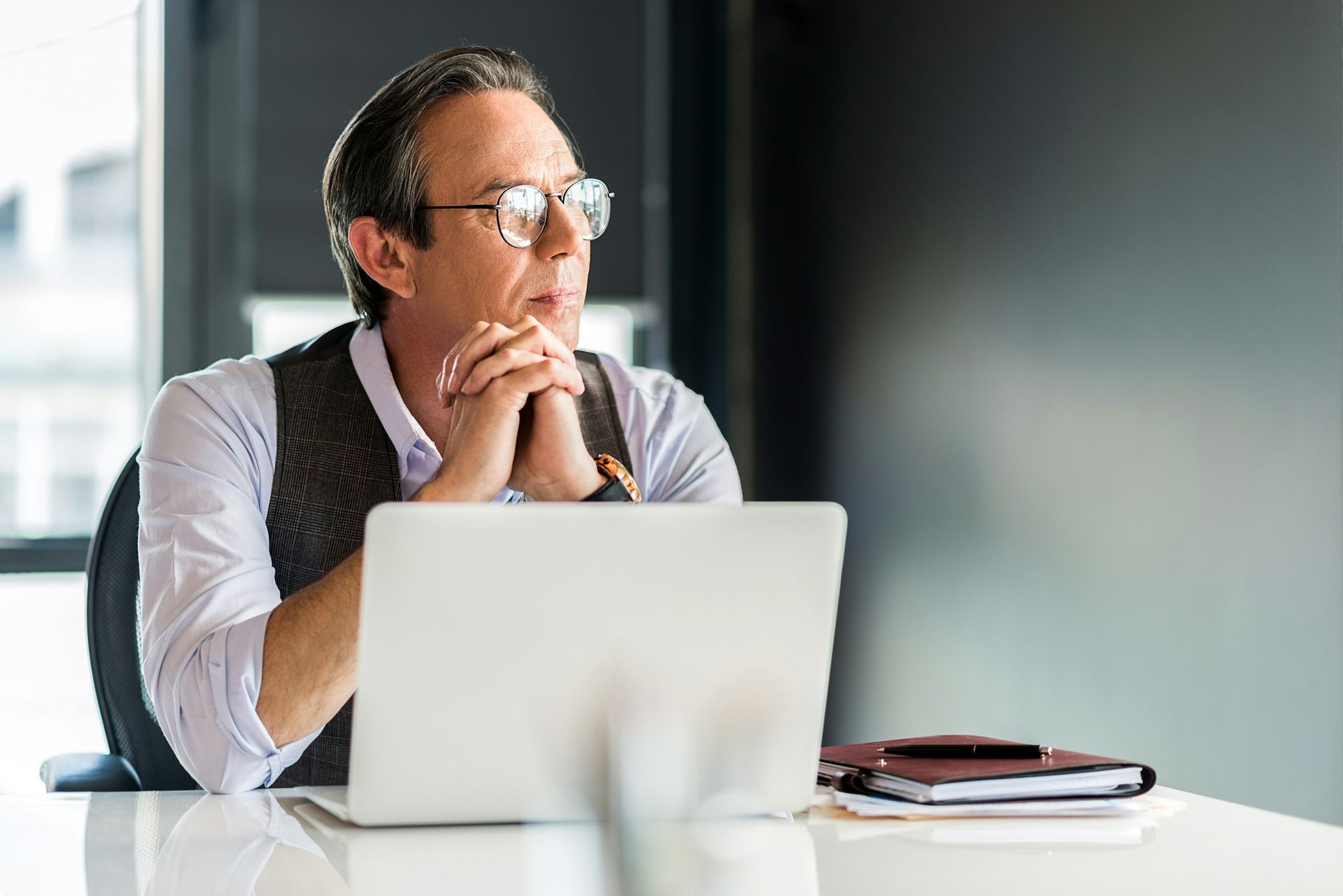 Man in glasses at desk, looking thoughtful, hands clasped, laptop open.