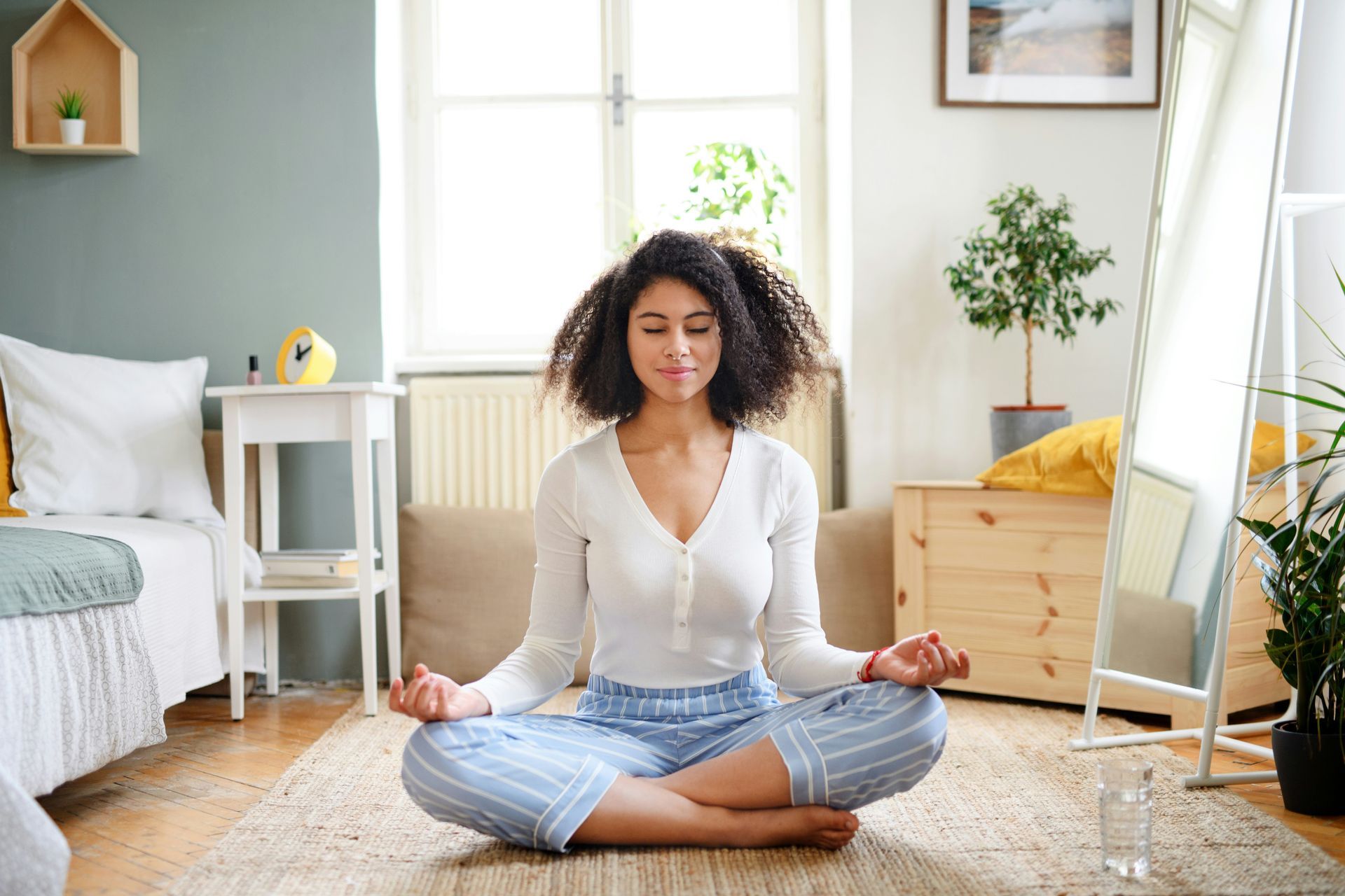 Woman with curly hair meditating on a rug in a bright bedroom, eyes closed, in a lotus position.