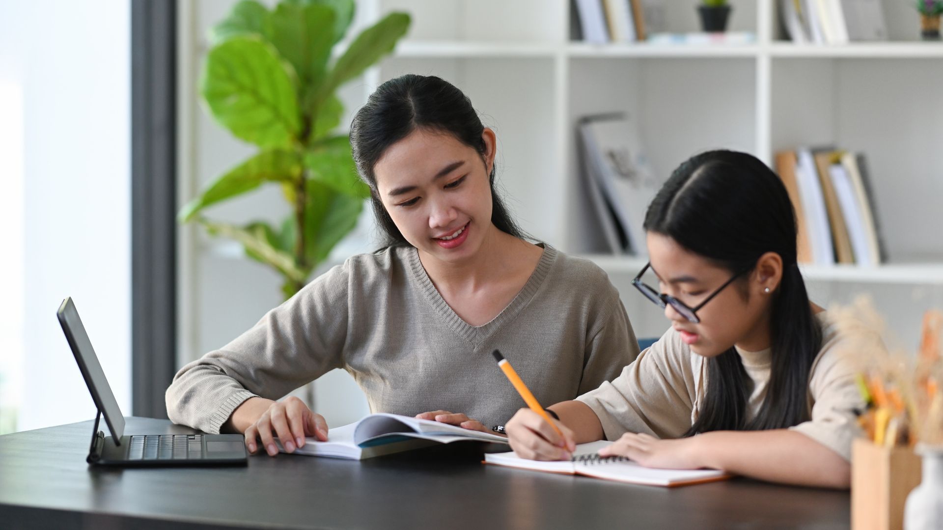 Woman assisting a girl with homework at a desk. They are smiling while reading a book and writing on paper, with a laptop nearby.