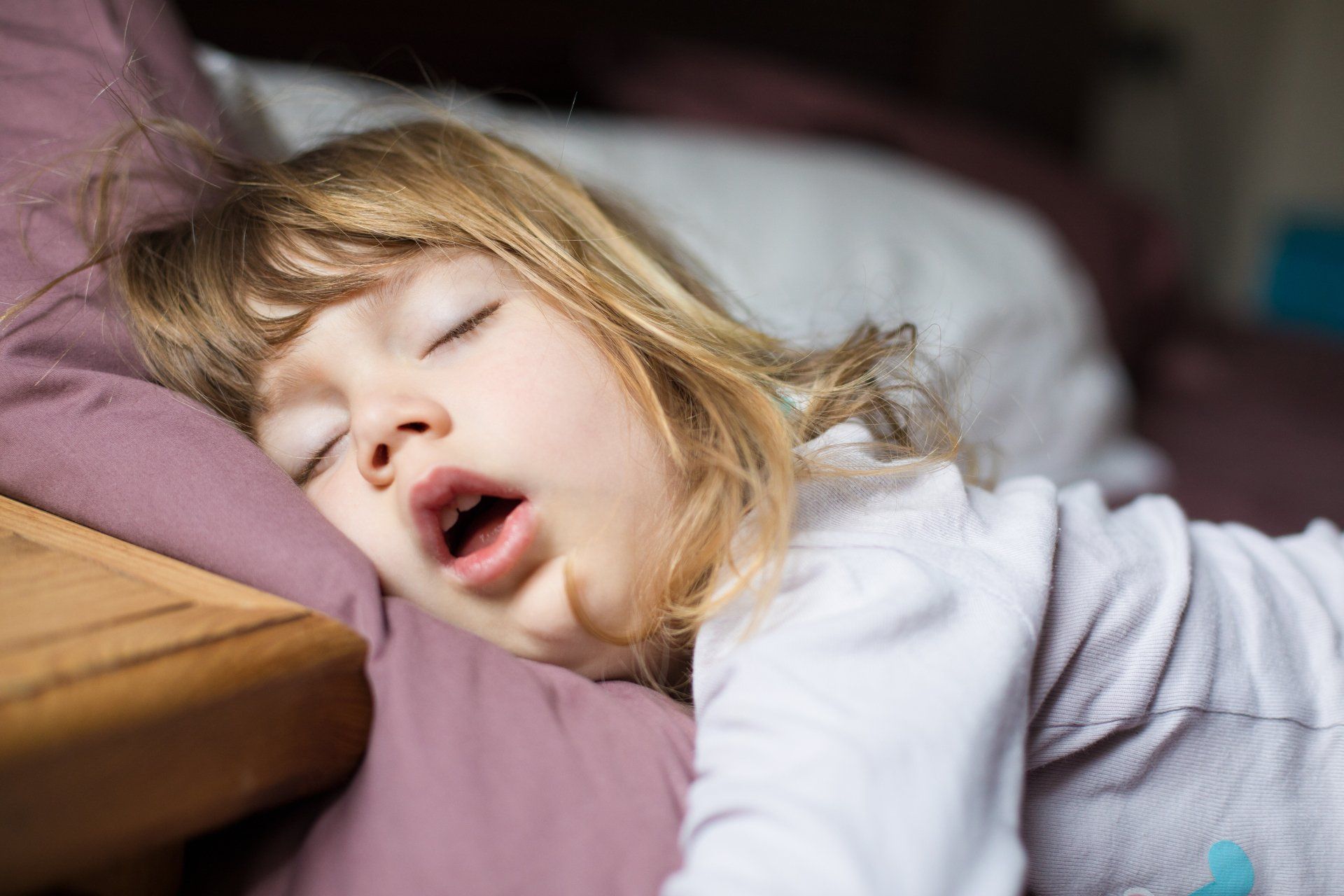Young child sleeping soundly on a bed, mouth open, blonde hair messy.