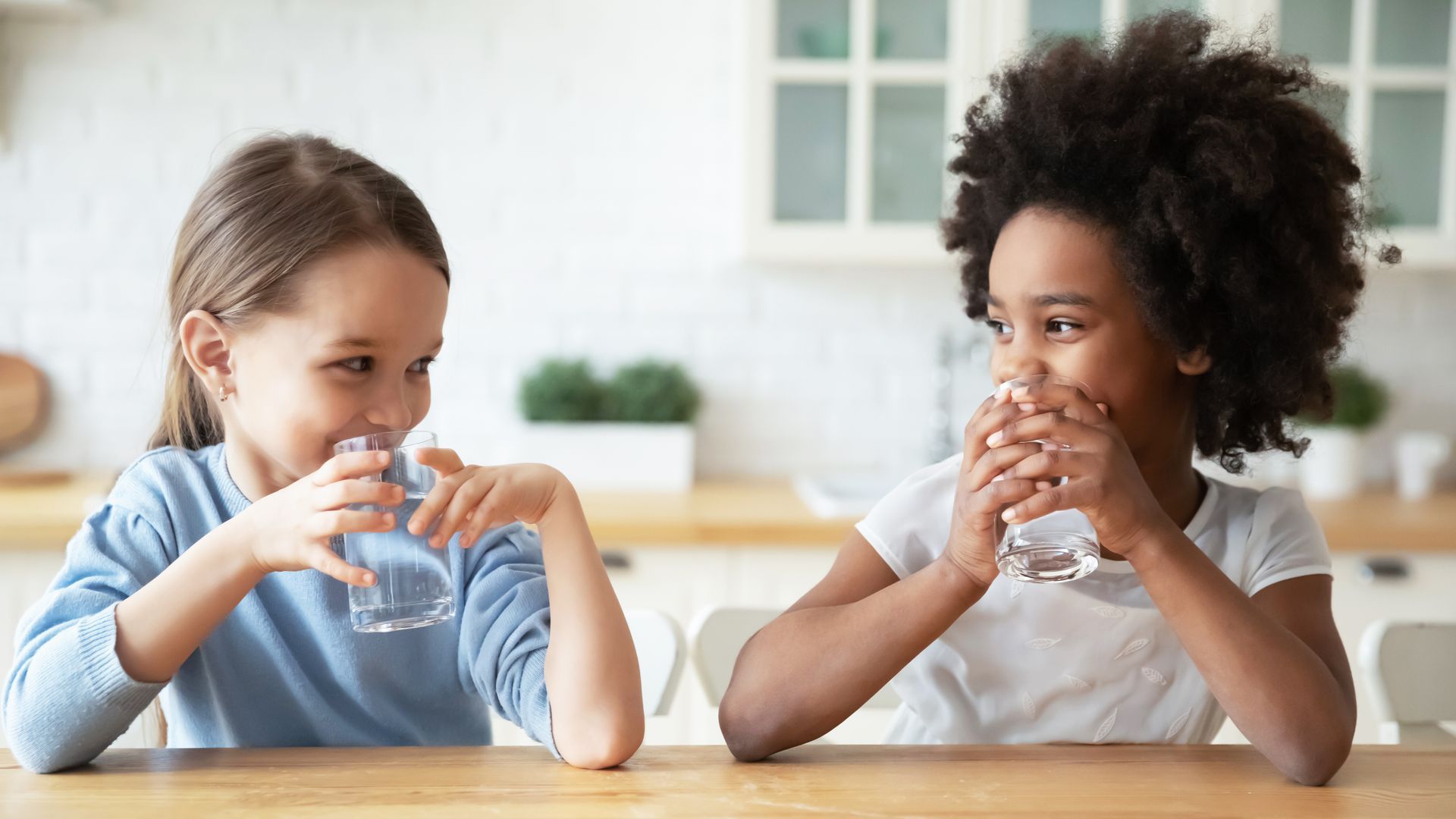 Two young girls drinking water at a wooden table in a kitchen. One has light skin, the other dark, both smiling.