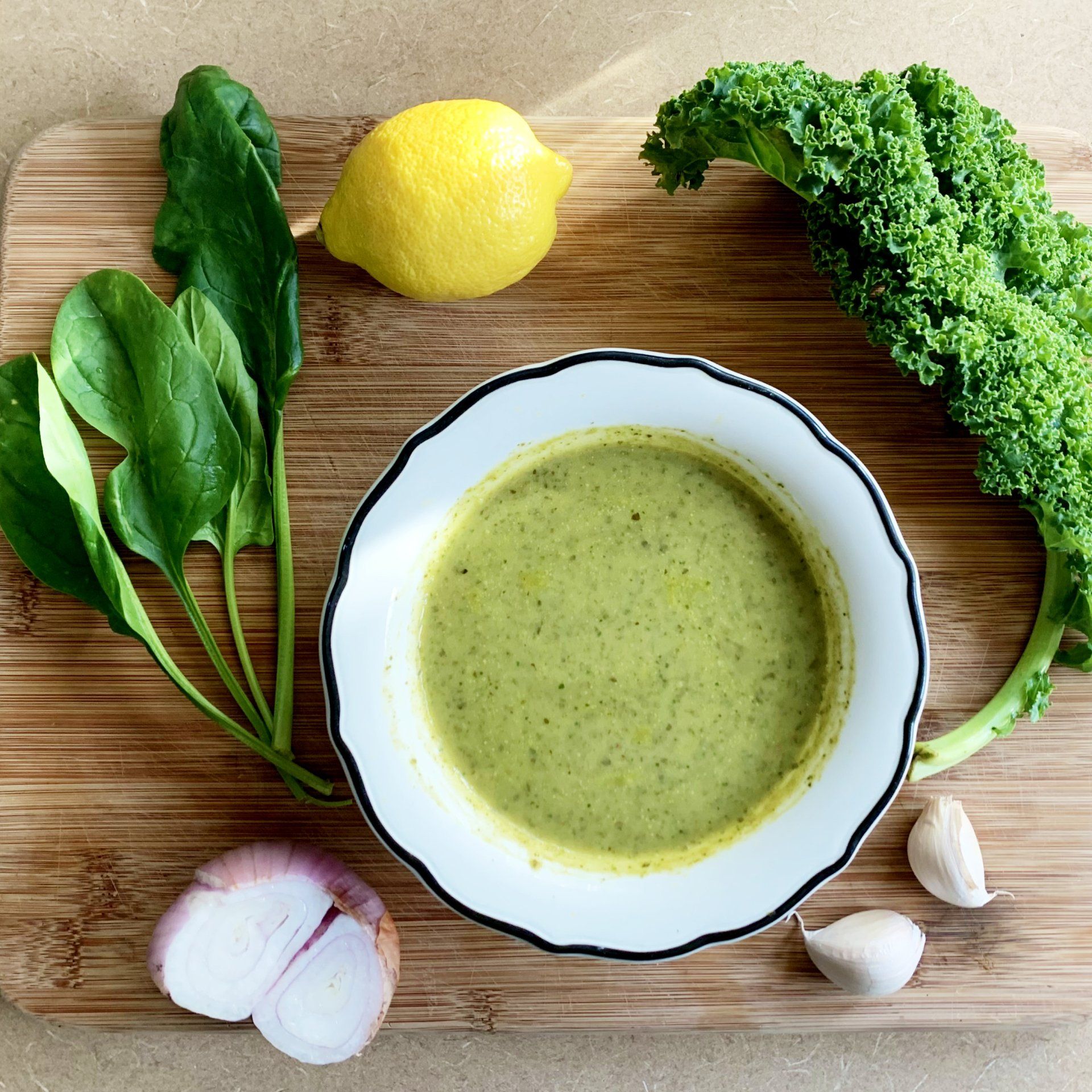 A bowl of green dressing surrounded by spinach, kale, lemon, shallot, and garlic on a wooden board.