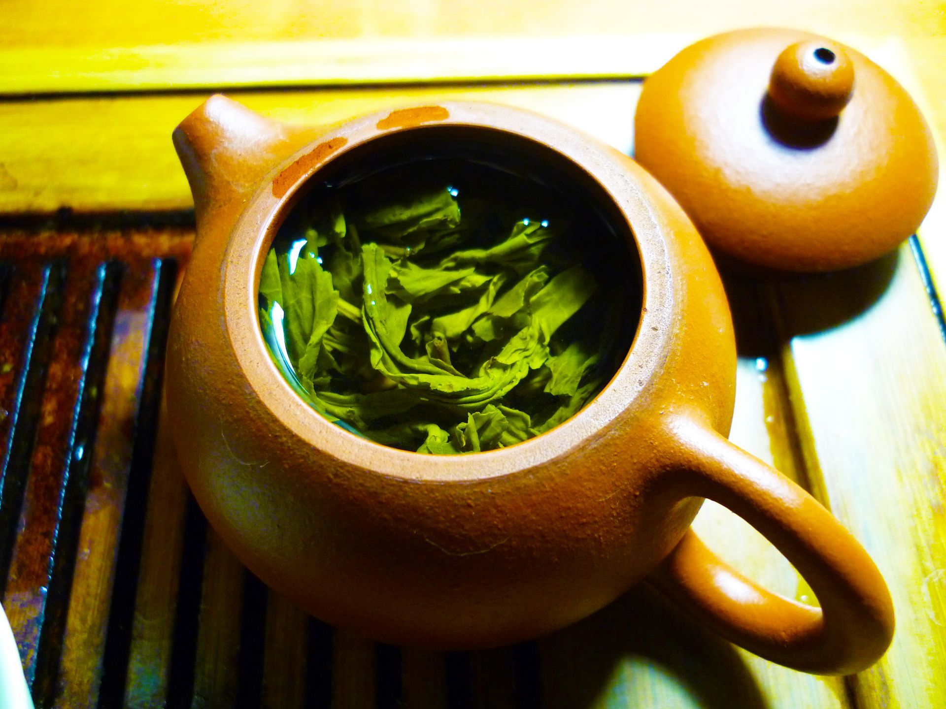 A small brown teapot filled with green tea leaves and hot water; the lid is beside it on a wooden tea tray.