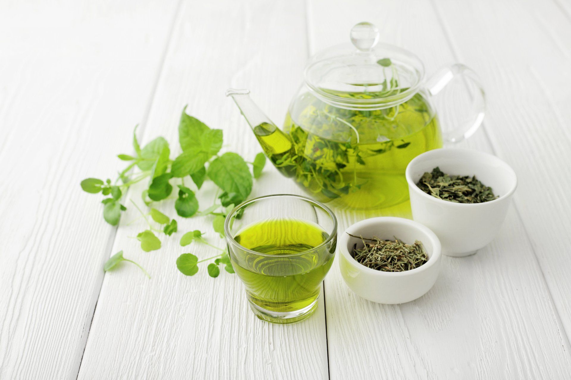 Green tea brewing setup: glass teapot, teacup, and two small bowls with tea leaves on a white wooden surface.