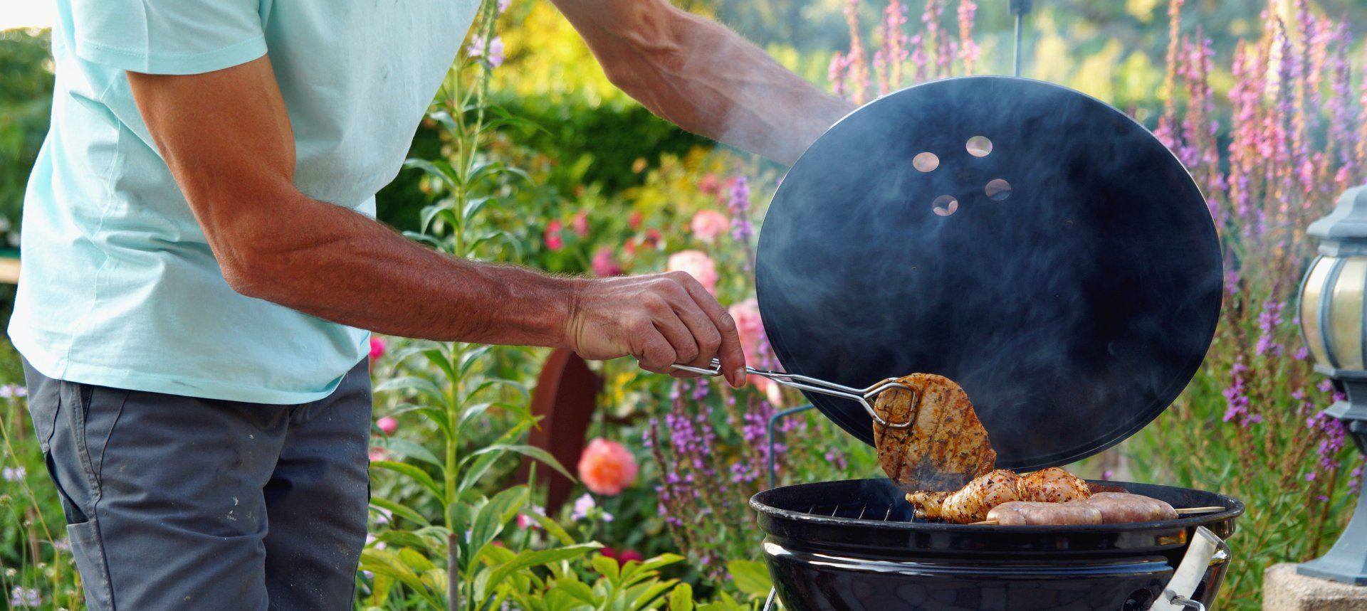 A person grilling outside, using a lid to scoop cooked food from a barbecue, with purple flowers in the background.