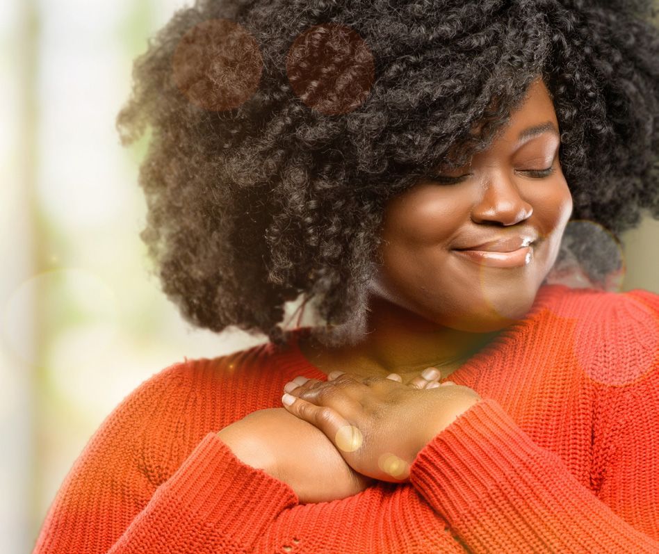 Smiling Black woman with curly hair, hands clasped over chest, wearing an orange sweater. Blurred background.