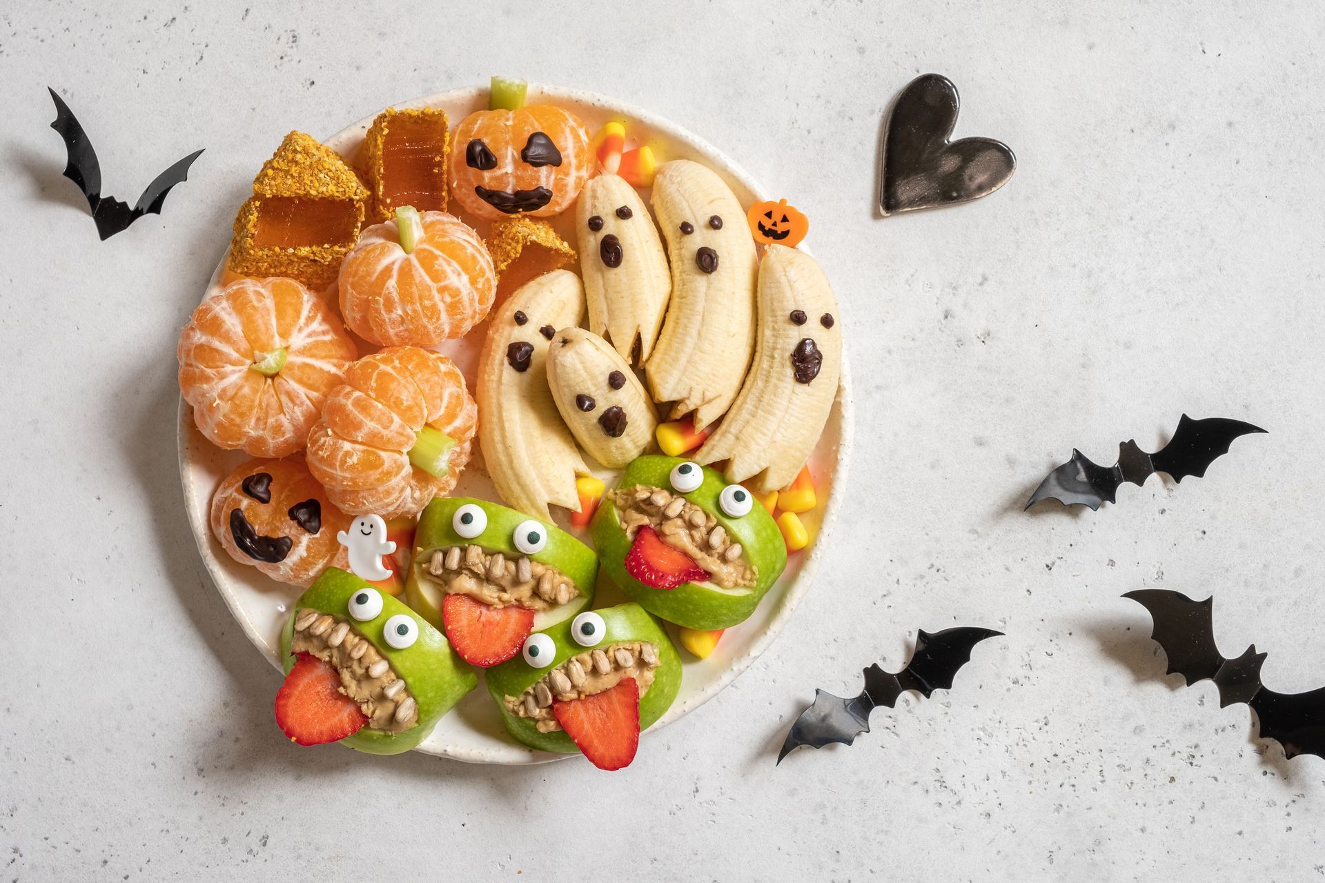 Halloween-themed fruit plate with mandarin oranges, banana ghosts, apple monsters, and pumpkin decorations on a white plate, surrounded by bat cutouts.