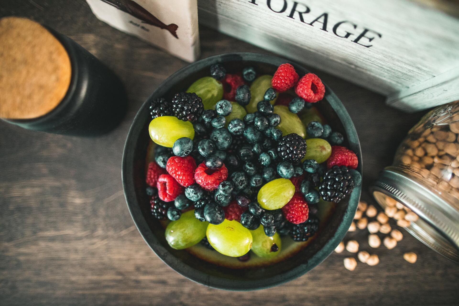 Bowl of fresh berries (blueberries, raspberries, blackberries, grapes) on a wooden surface, with pantry items in the background.