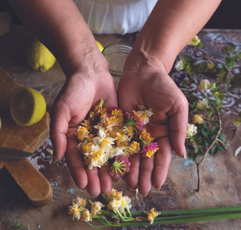 Woman's hands cupped, holding colorful flower petals.  Lemons and a cutting board are visible on the rustic table.