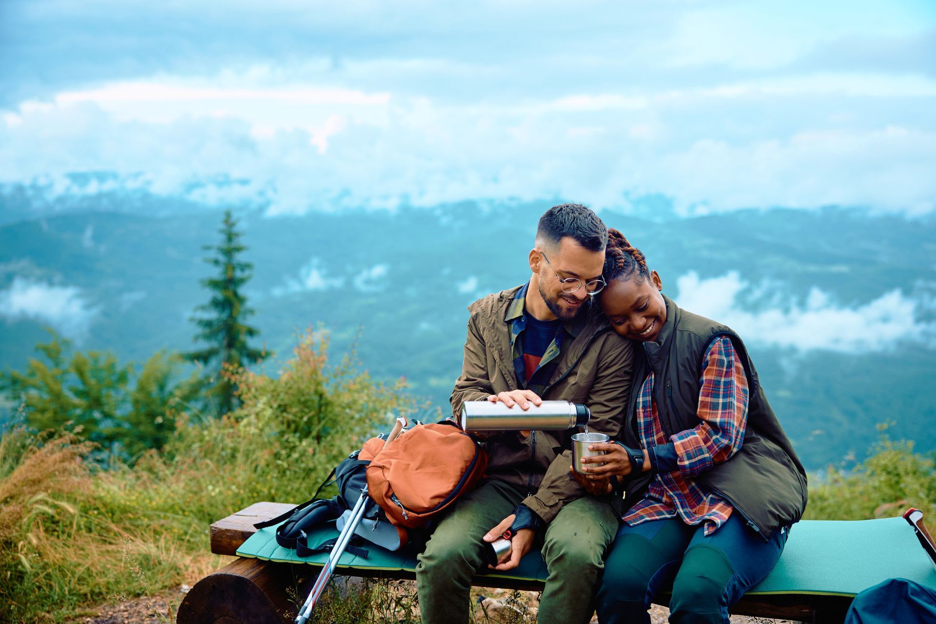 Couple on a mountaintop, drinking from a thermos. Man pours for woman who leans on his shoulder. Greenery and misty mountains in the background.