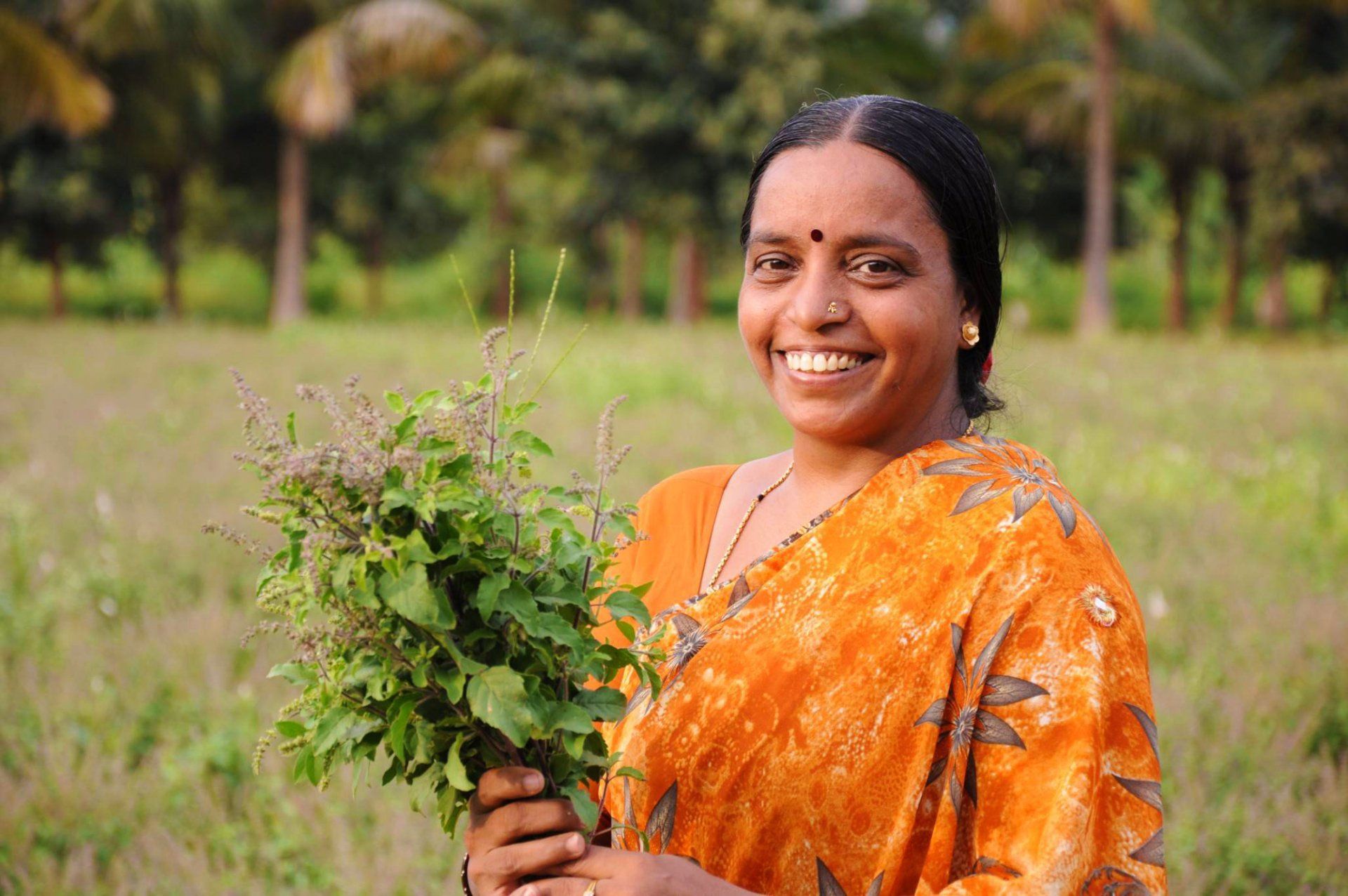 Woman in orange sari smiling, holding basil in a field. Background trees and green grass.