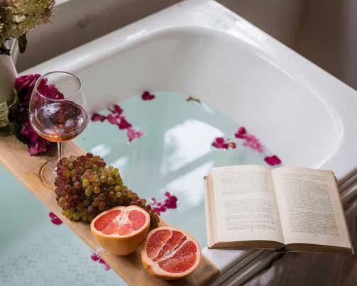 A bathtub with pink flower petals floating in the water, a book, and a tray holding grapes, grapefruit, and a glass of wine.