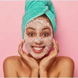 Woman with a facial scrub and a green towel on her head, smiling with hands near face, against a pink background.