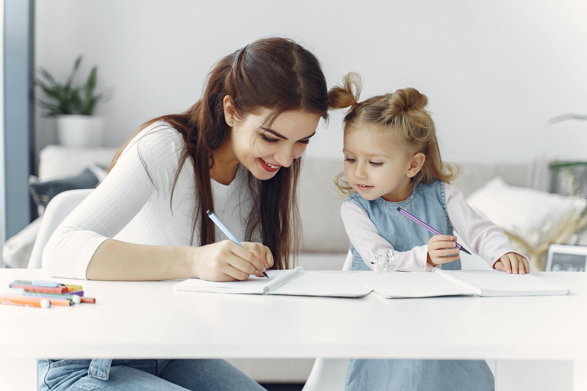 Woman and young girl writing at a white table indoors. The woman smiles, and the girl looks on with interest.