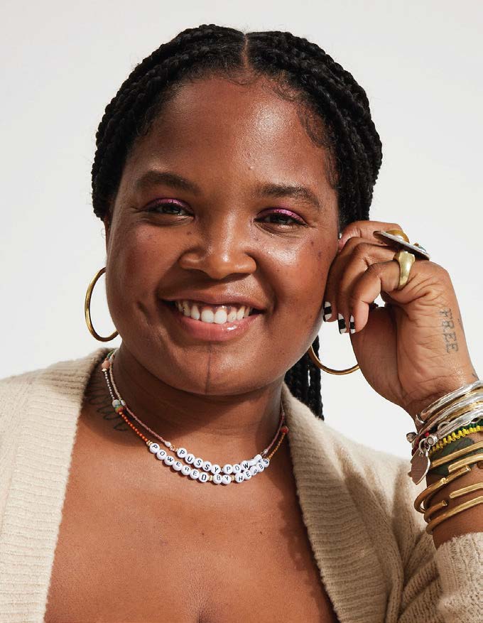 Woman with braided hair smiles broadly, wearing hoop earrings, a beaded necklace, and many bracelets. She has pink eyeshadow and is in a neutral-toned setting.