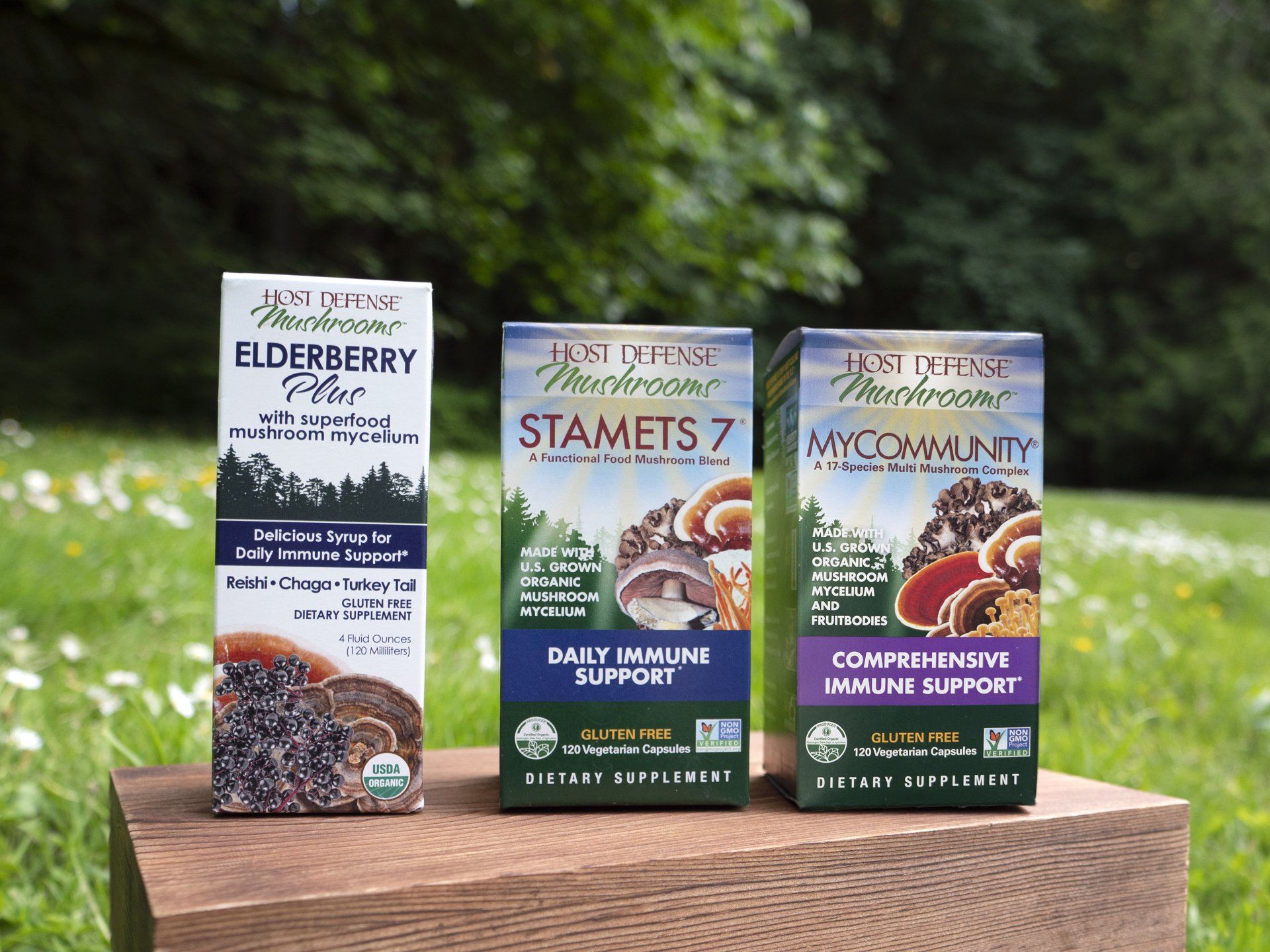 Three boxes of Host Defense mushroom supplements on a wooden surface, against a blurred green outdoor background.