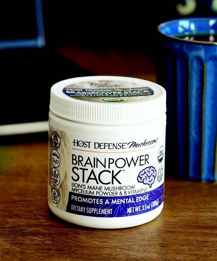 White jar of Host Defense Brain Power Stack supplement on a wooden table with a blue mug in the background.