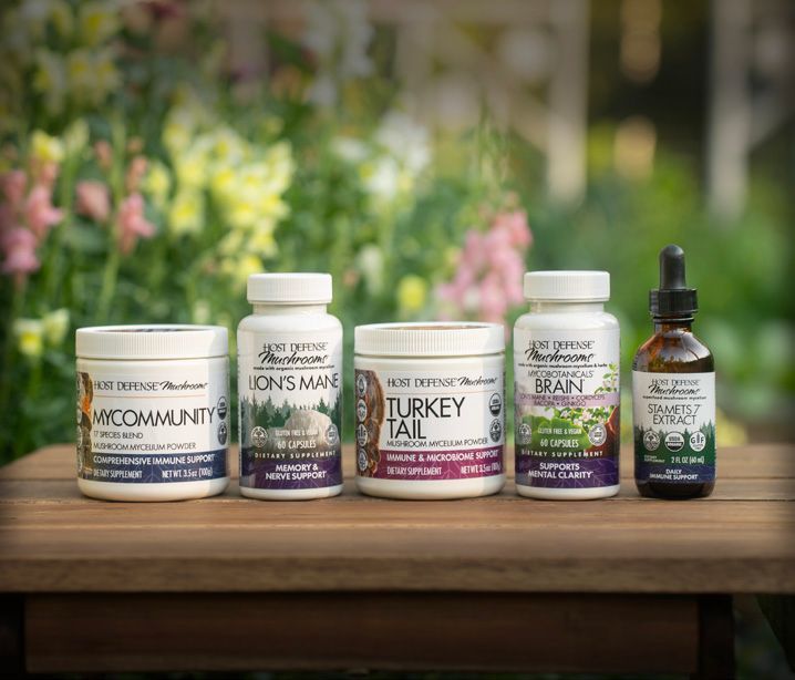 Five mushroom supplements on a wooden table outdoors in front of a blurred floral background. Various labeled jars and a dropper bottle are displayed.