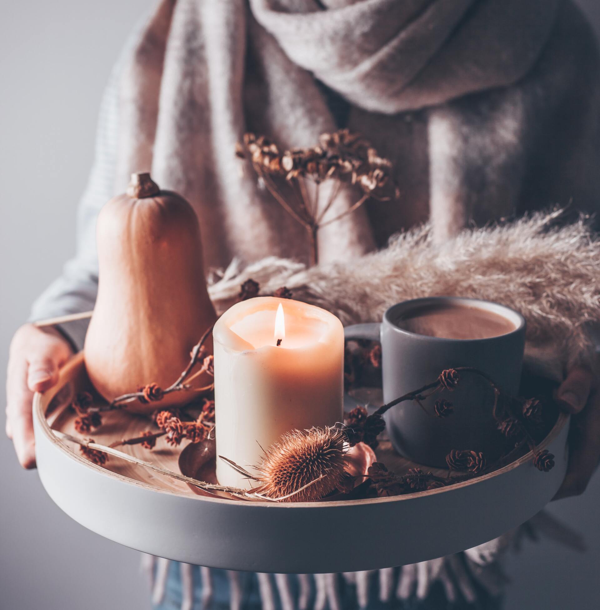 Person holding a tray with a lit candle, gourd, coffee, and dried floral decorations, in warm tones.
