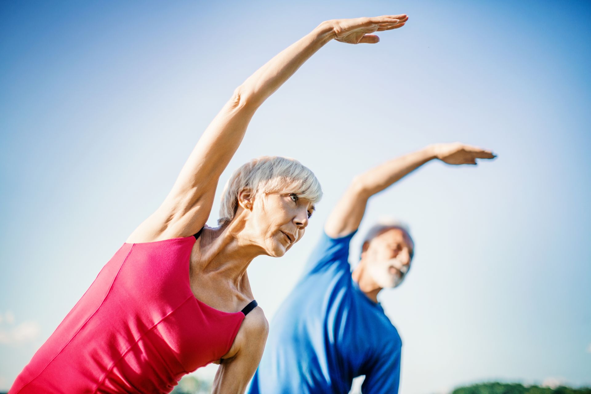 An older woman in a pink top and an older man in a blue shirt stretch outdoors under a blue sky.