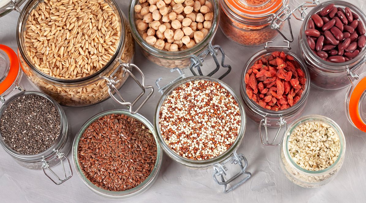 Glass jars filled with various grains and legumes: oats, chickpeas, lentils, red beans, goji berries, quinoa, chia seeds, flax seeds, and brown rice.