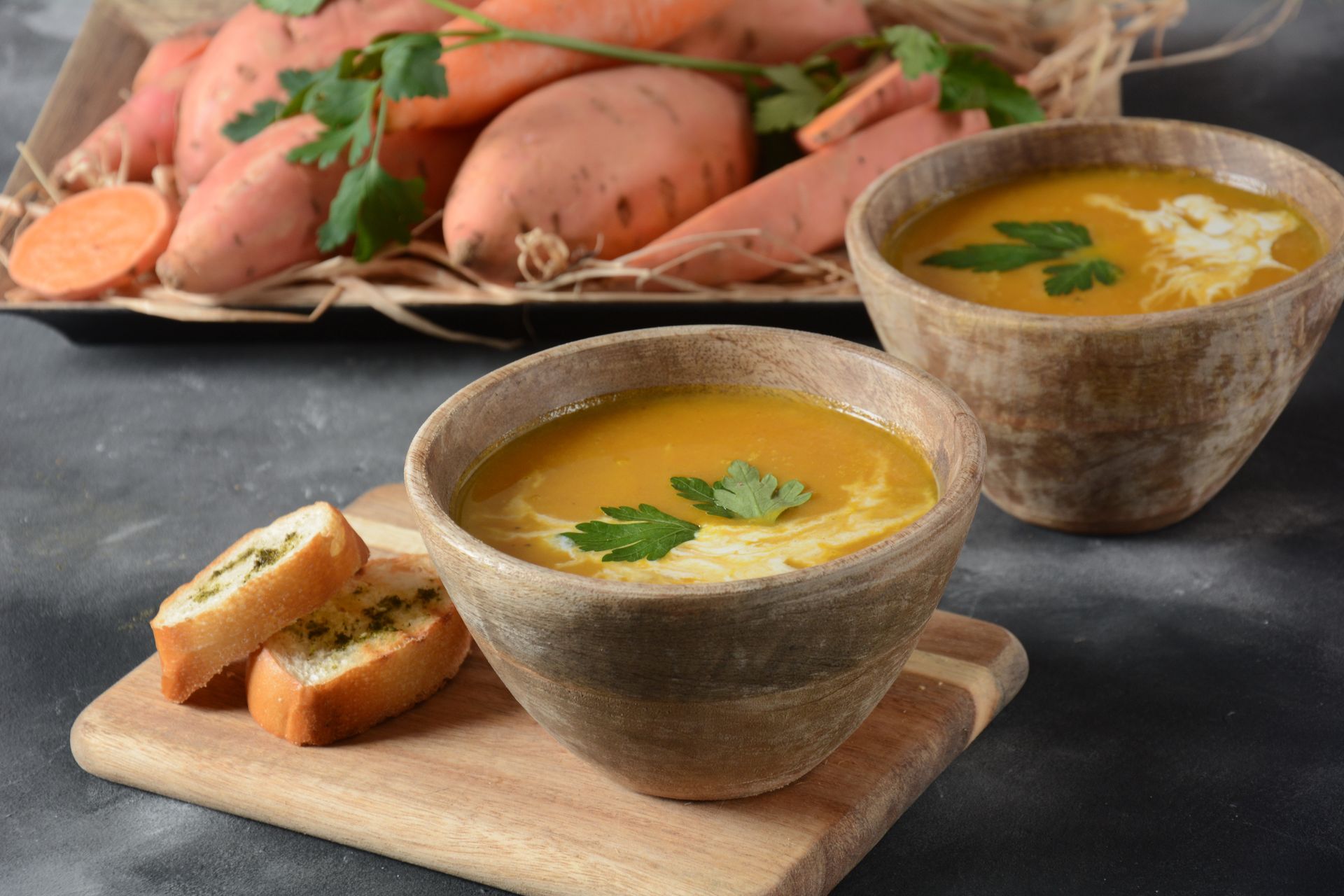 Two bowls of orange sweet potato soup with bread and sweet potatoes in the background.
