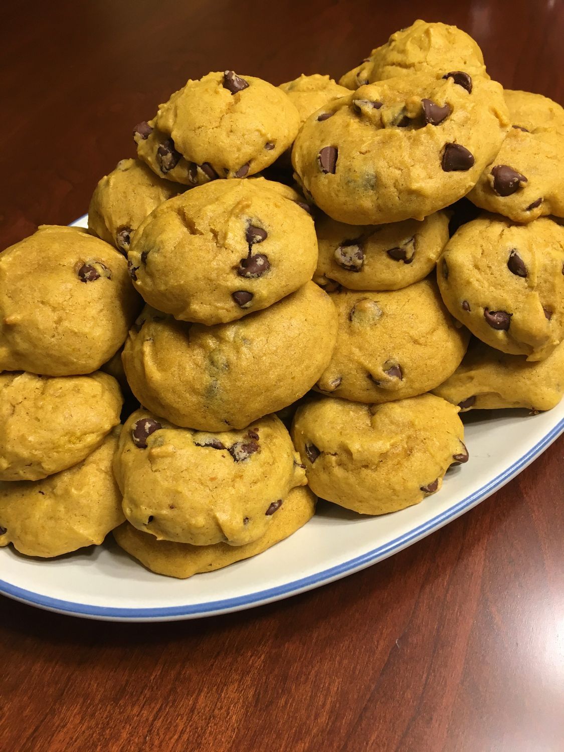 Pile of pumpkin chocolate chip cookies on a white plate with a blue rim, on a dark wooden table.