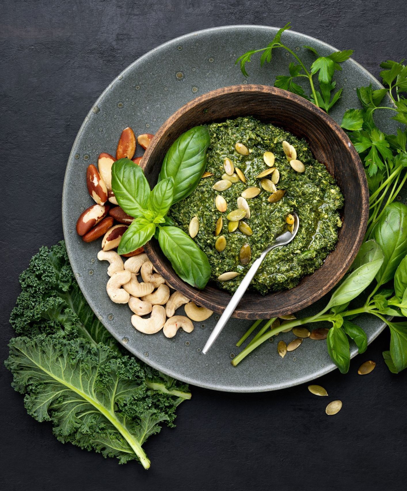 Green pesto in a wooden bowl with basil leaves and pumpkin seeds, served with nuts and kale on a gray plate.