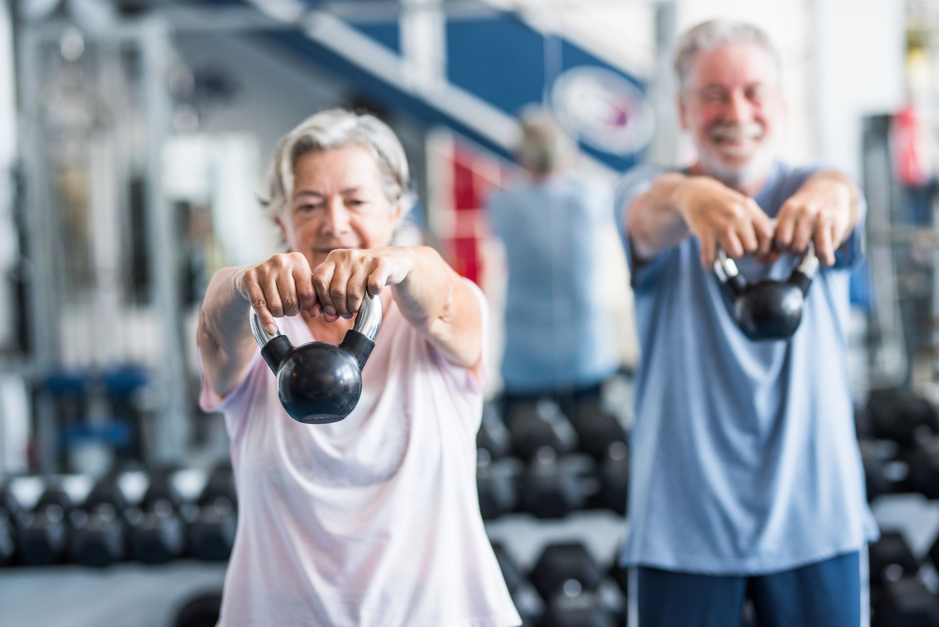 Two older adults working out with kettlebells at a gym. They are smiling and extending their arms forward.