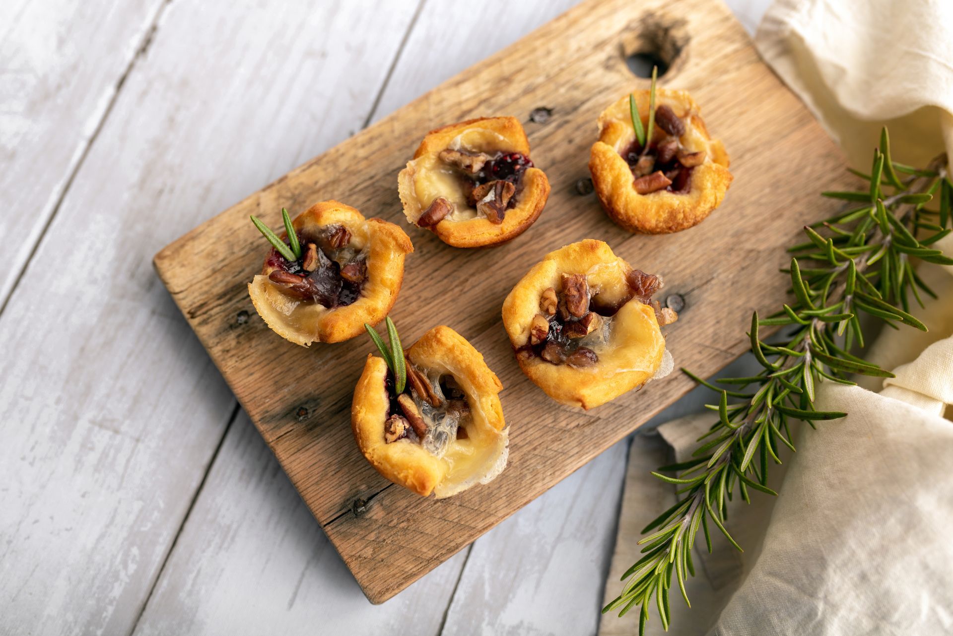 Mini tarts with a pecan topping sit on a wooden board with sprigs of rosemary.