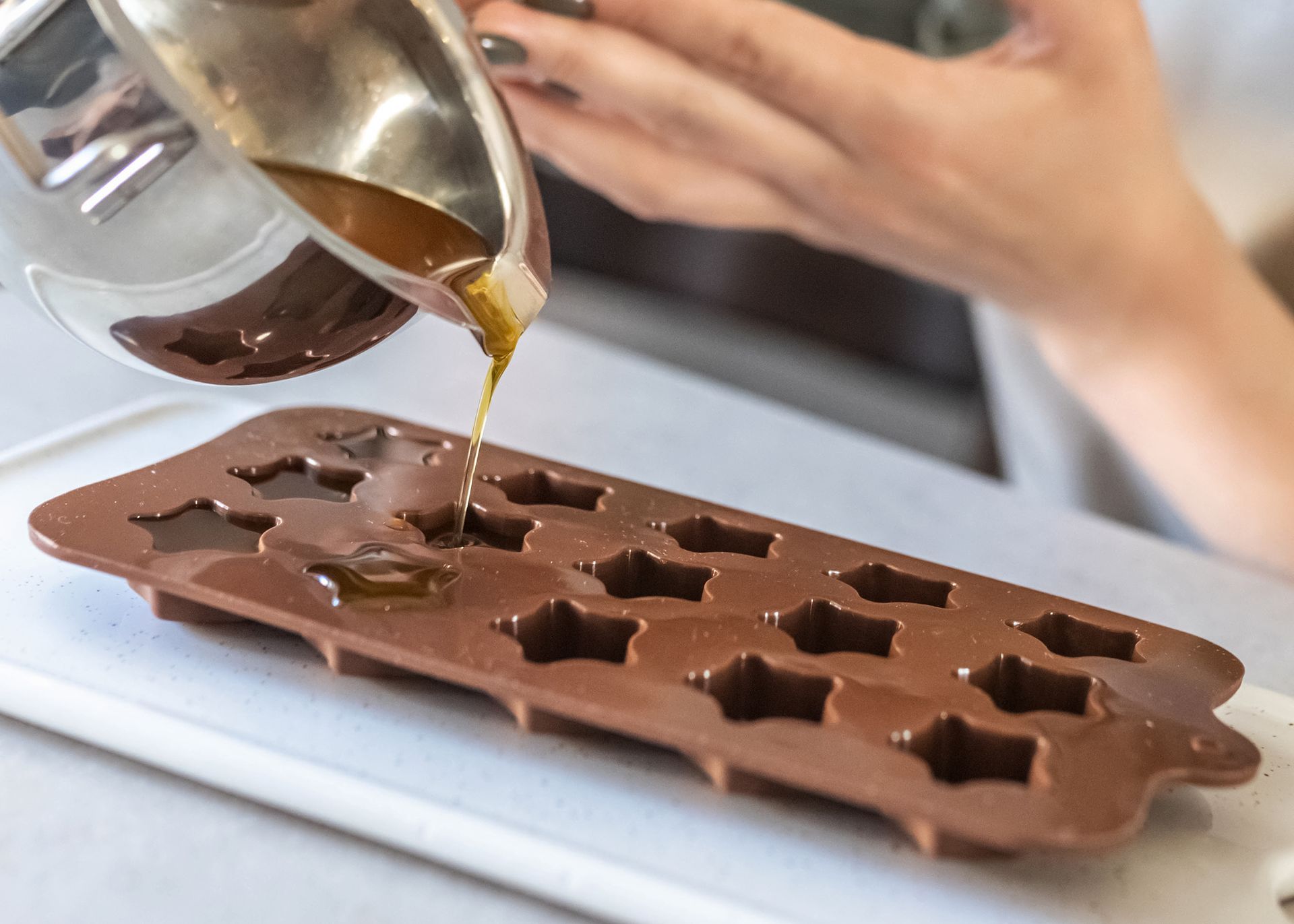 Brown liquid is being poured from a metal pot into a brown star-shaped silicone mold. A person's hands are visible.