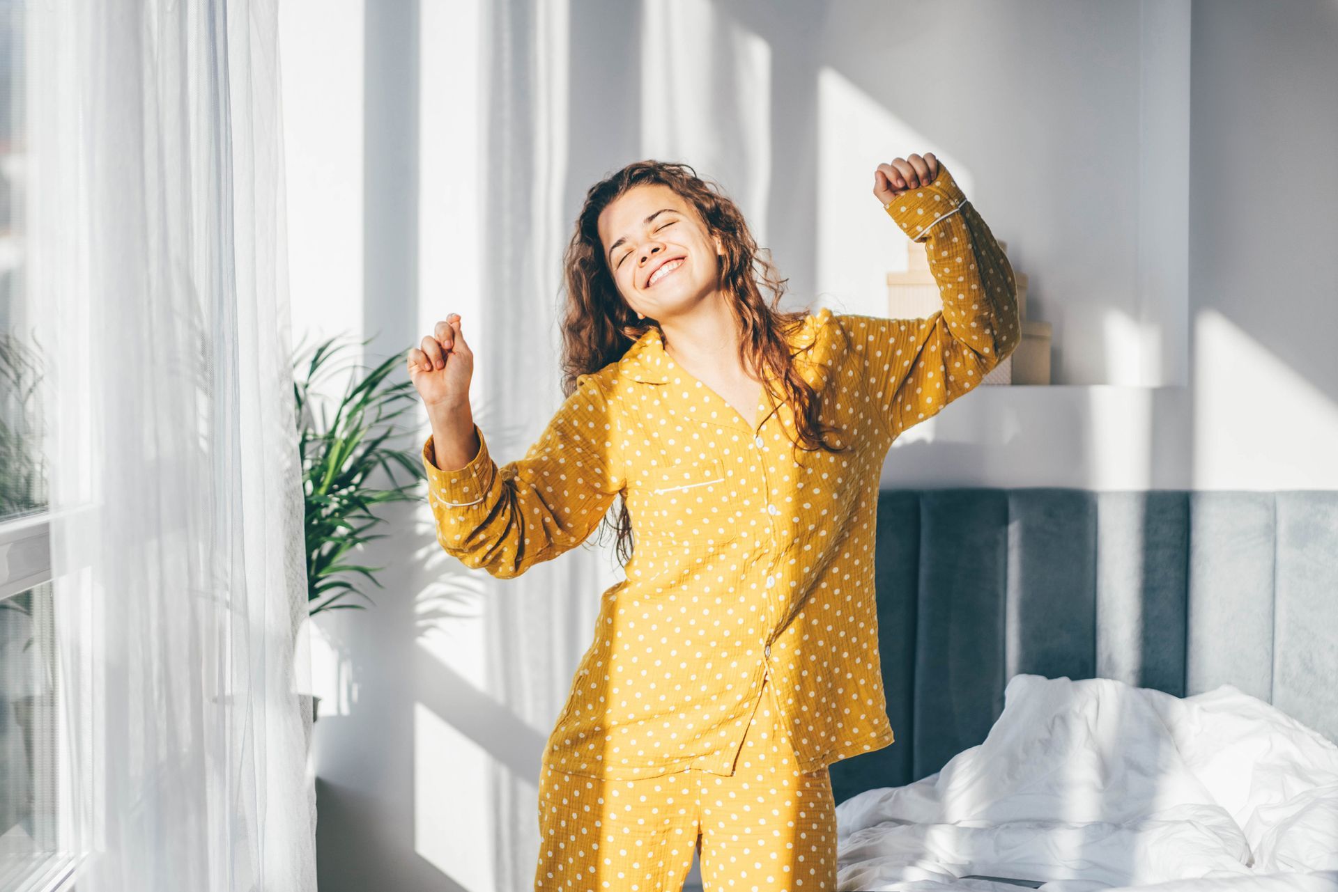 Woman in yellow polka dot pajamas stretching in a sunny bedroom, with closed eyes and a smile.