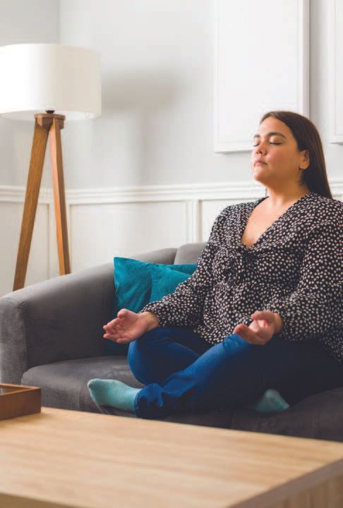 Woman meditating on a couch in a living room, eyes closed, hands in a yoga pose, wearing blue jeans and a patterned blouse.