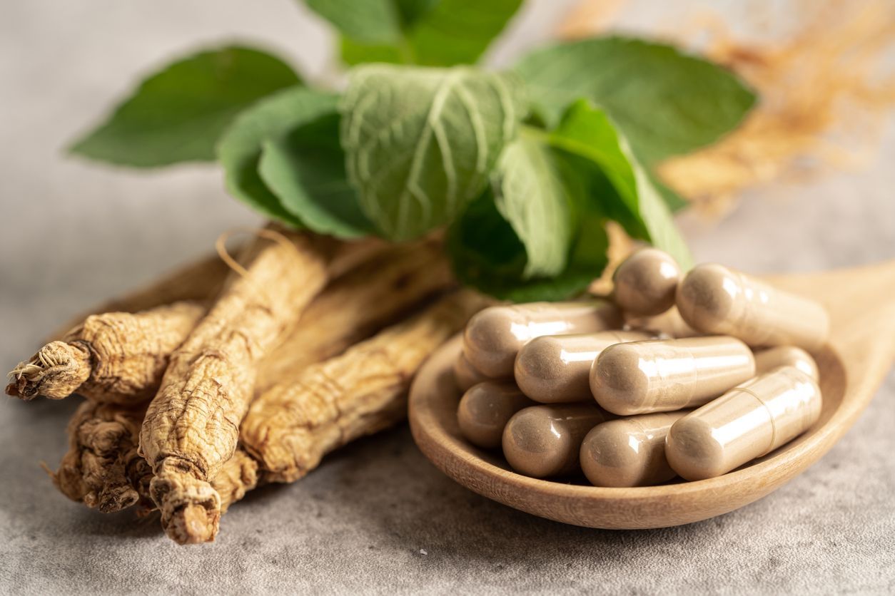 Ginseng roots, fresh green leaves, and brown capsules rest on a wooden spoon, set on a gray surface.